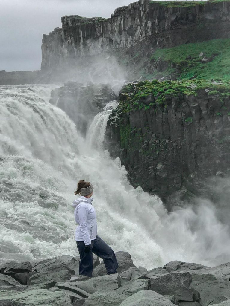 girl in a white jacket standing in front of a gushing waterfall