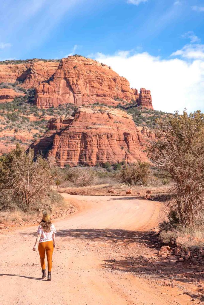 A girl walking down a red dirt pathway towards a field of towering red-rock cliffs.
