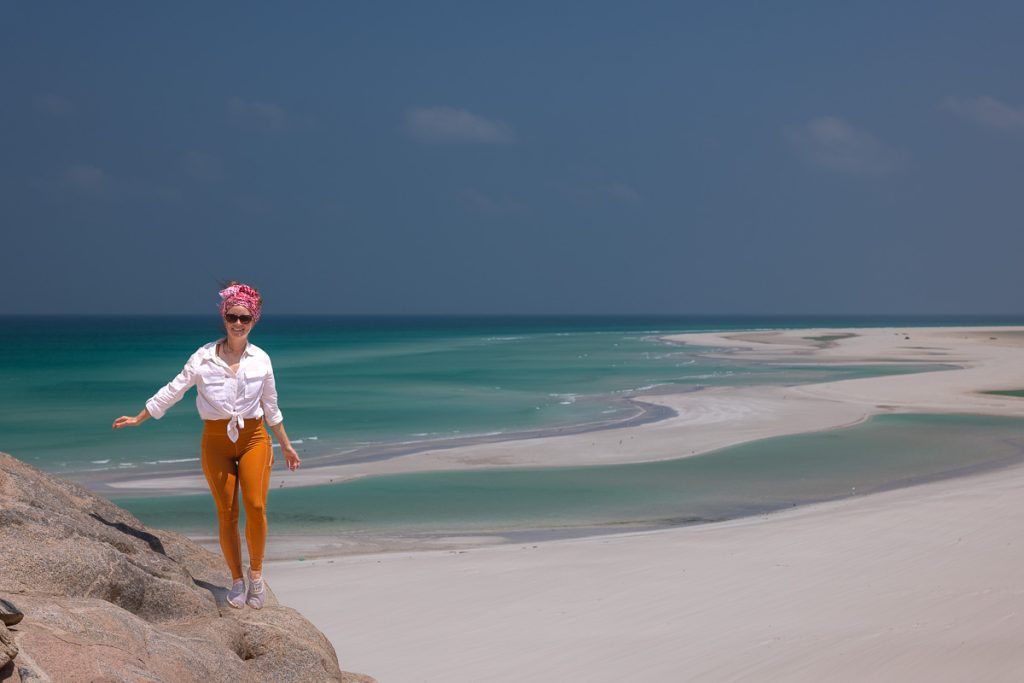 Sam in a white top and orange pants walking along a rock ledge overlooking aqua water and white sandy beach.