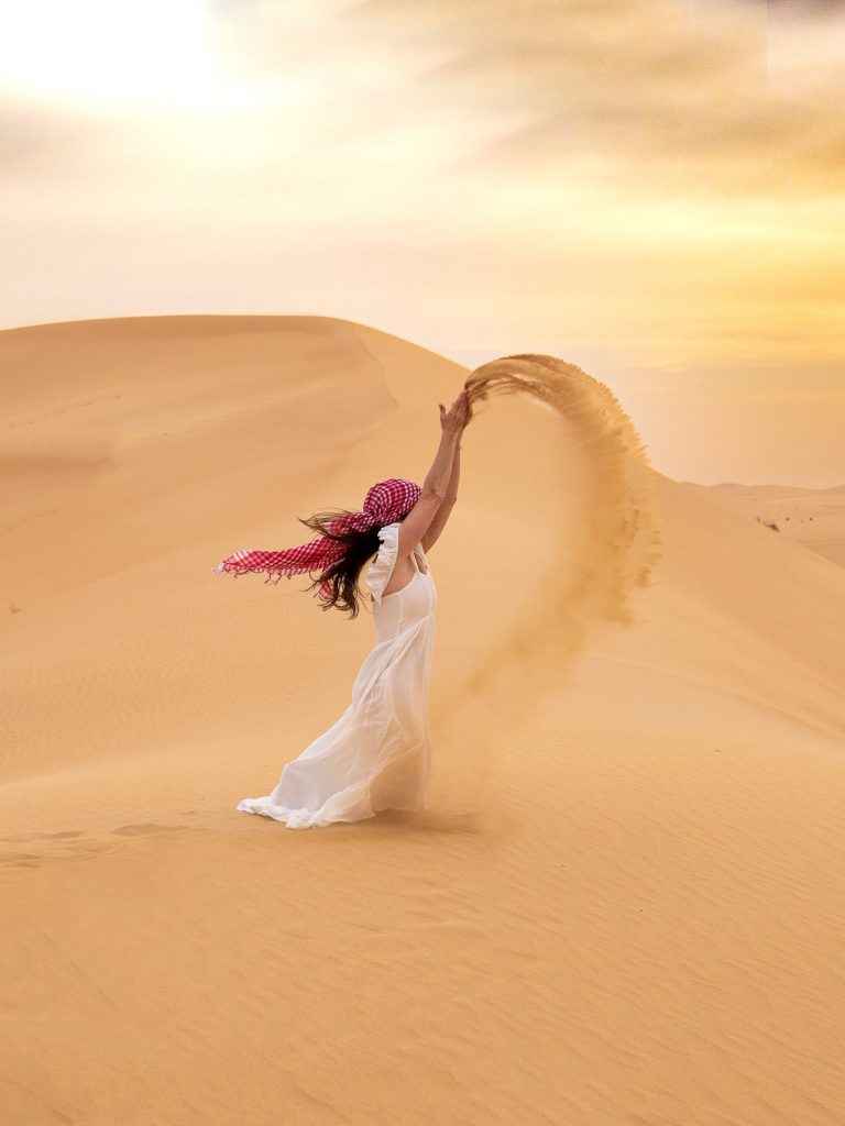 Sam in a white dress throwing sand above her head in the desert.
