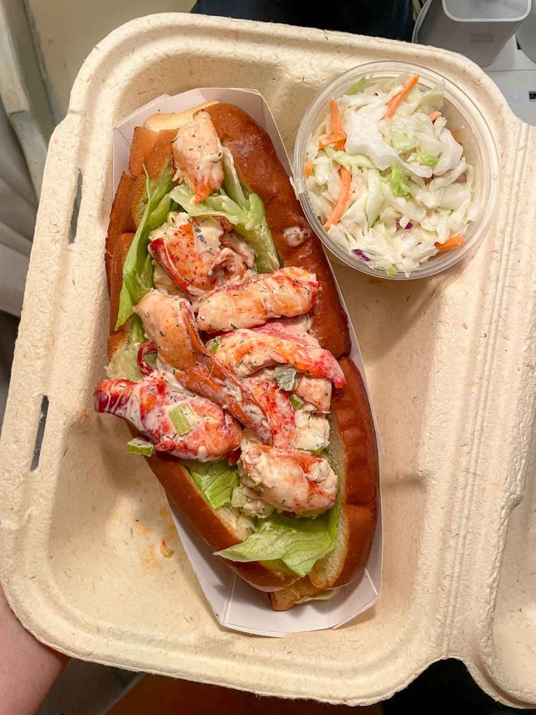 Dressed lobster roll in a carton to go container with a side of cole slaw.