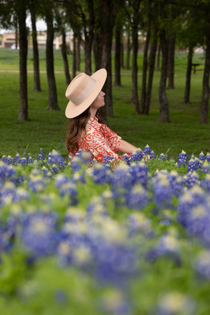 Girl in a hat sitting behind purple flowers.